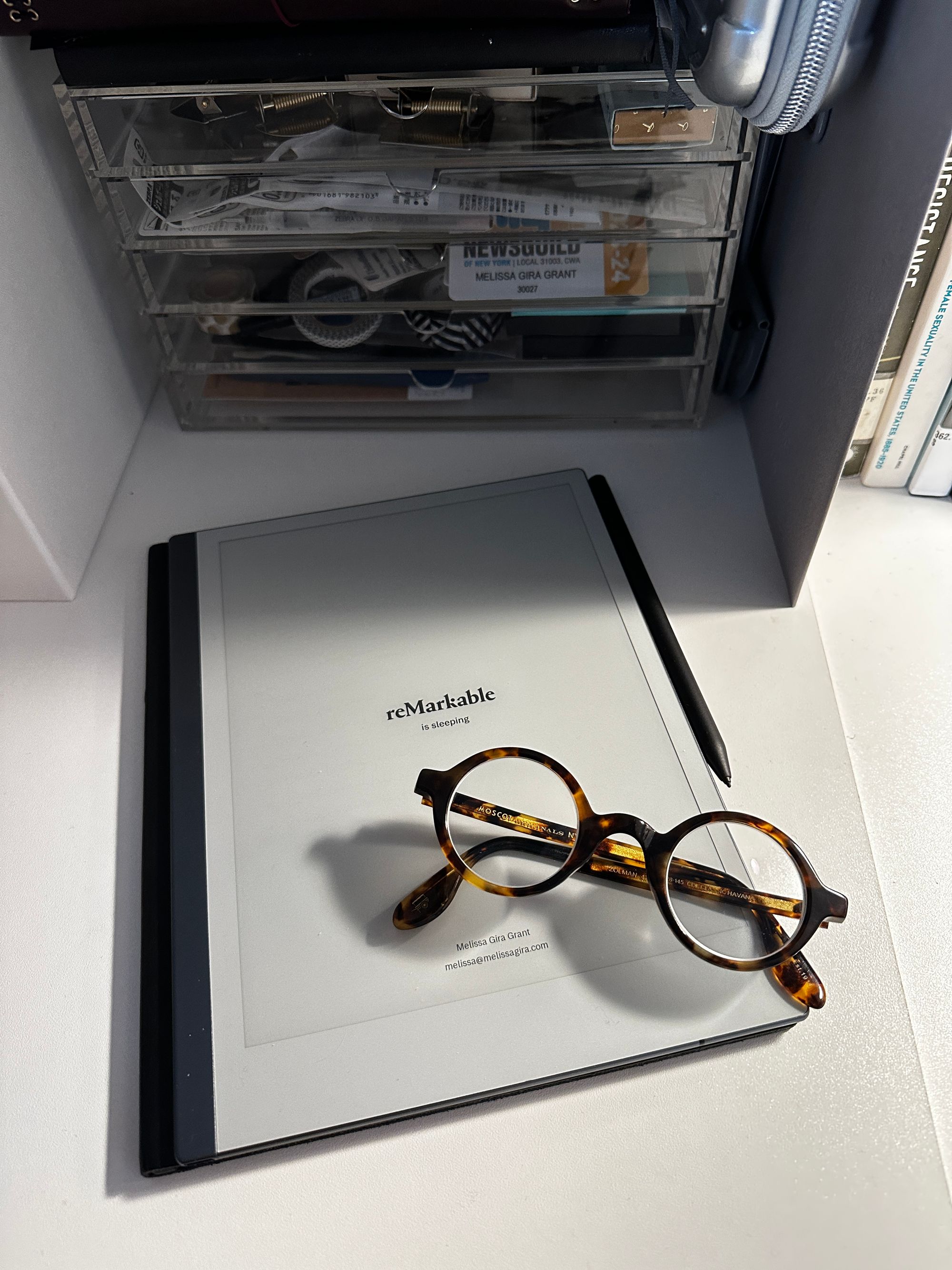 My reMarkable tablet and my round tortoiseshell glasses on my white desk top with an acrylic mini drawer (in which you can see some washi tape, my NewsGuild card, some bronze clips) 